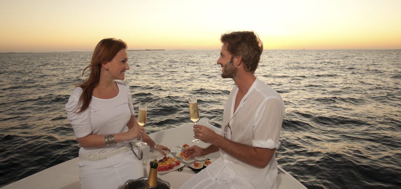 Un couple souriant avec des flûtes de Champagne sur un bateau au coucher du soleil sur l'eau.